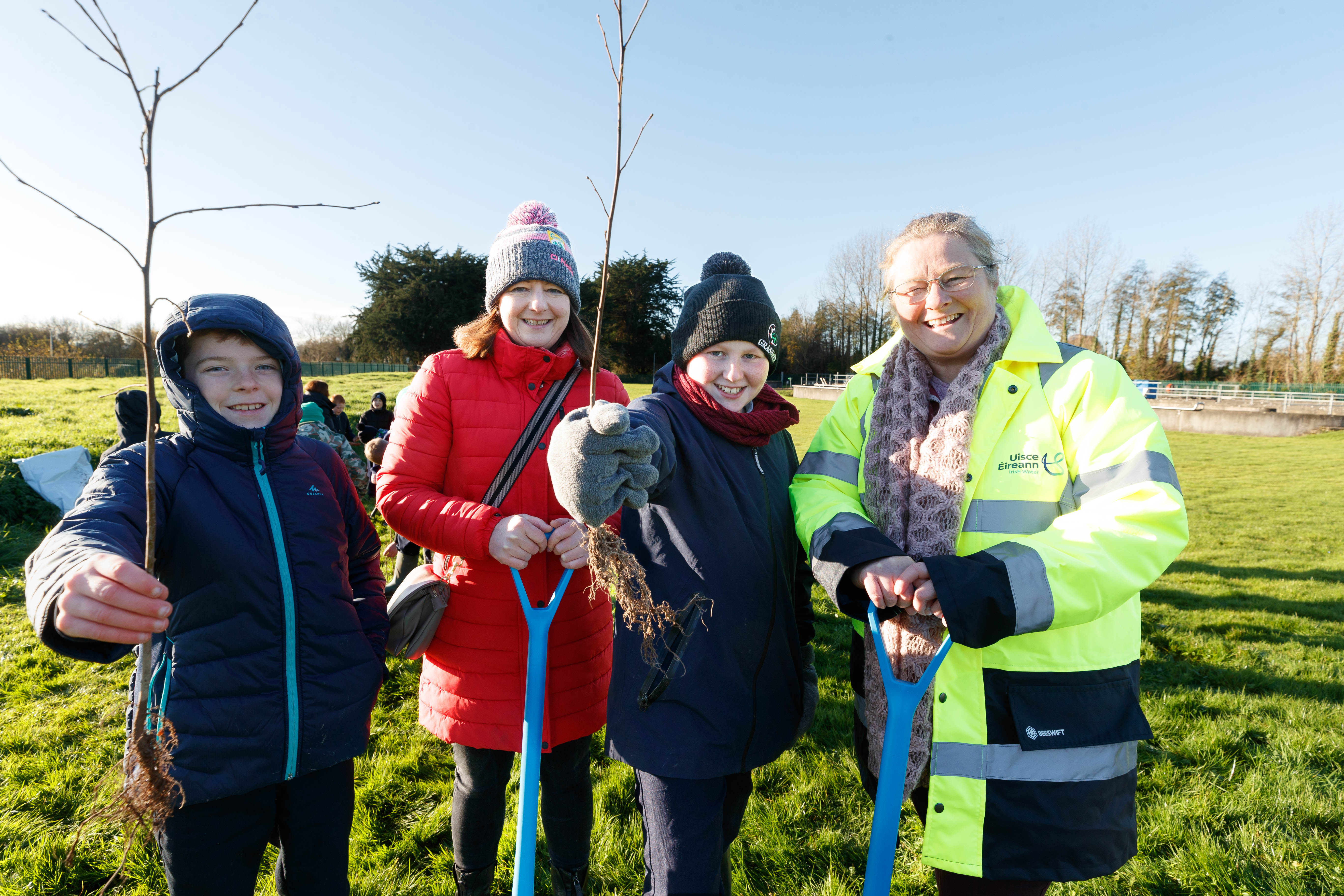 A group of two kids holding small trees to plant and two adults holding shovels