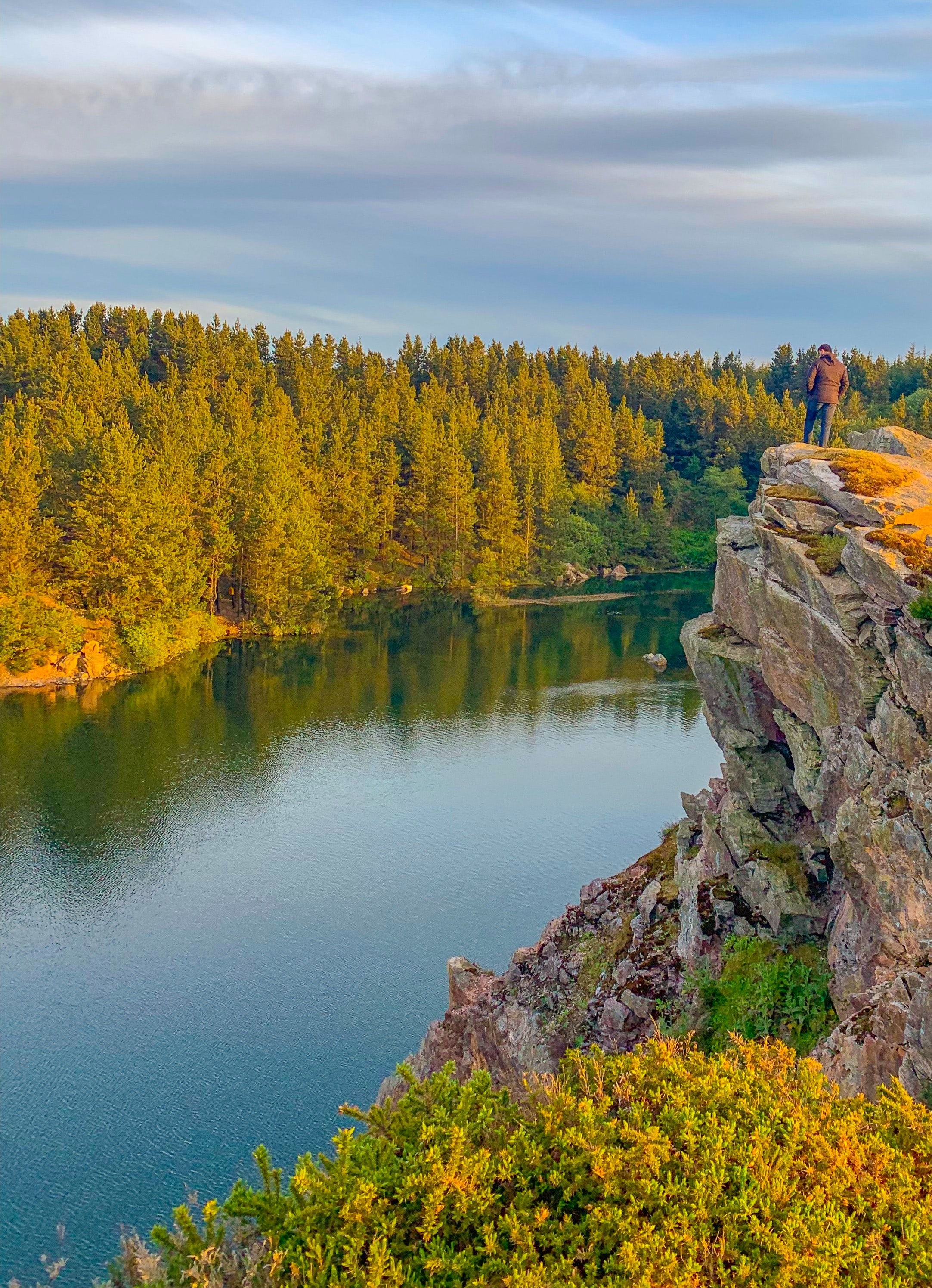 A man standing on a cliff above a large lake beside a forest